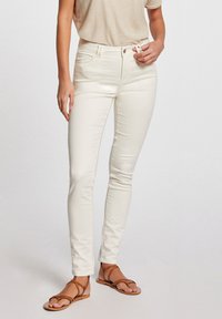 Jeans ajustados blancos con una textura suave, cintura media y costuras decorativas en los bolsillos, combinados con sandalias planas marrones con tiras.