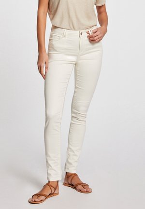 White slim-fit jeans with a smooth texture, mid-rise waist, and decorative stitching on pockets, paired with brown flat strappy sandals.