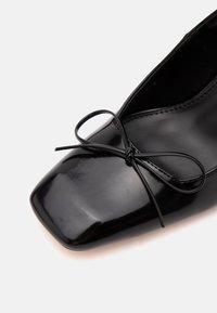 Gros plan d'une chaussure plate en cuir verni noir avec un bout carré et un petit nœud fin noué sur le dessus.