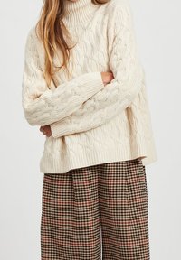 Pull à col montant en tricot torsadé crème avec des poignets côtelés, associé à un pantalon large à carreaux aux teintes marron, rouge et noir.