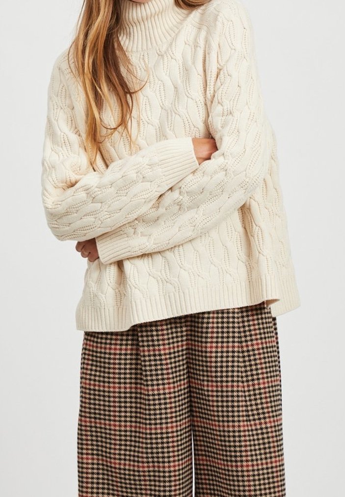 Pull à col montant en tricot torsadé crème avec des poignets côtelés, associé à un pantalon large à carreaux aux teintes marron, rouge et noir.