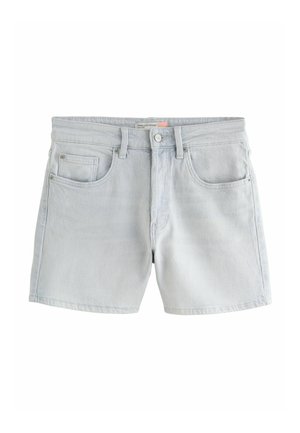 Nő sárga kötött pulóverben, banán mintákkal és világosszürke rövidnadrágban, egy struktúrált kőfalnak támaszkodva.