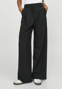 Zwarte wijde broek van een glad materiaal. Kenmerkt zich door een hoge taille, plooien aan de voorkant en zijzakken. Gecombineerd met witte sneakers.