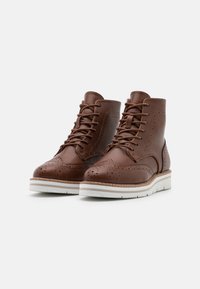 Bottines en cuir marron avec détails en brogue, design à lacets, semelle blanche contrastante et accents cousus le long des bords.