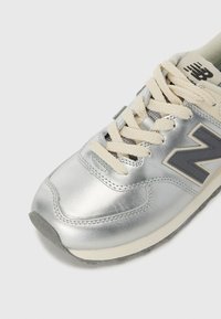 Zapatilla plateada con un exterior metálico suave, cordones beige y un logo gris en el costado, con una suela texturizada para mayor agarre.