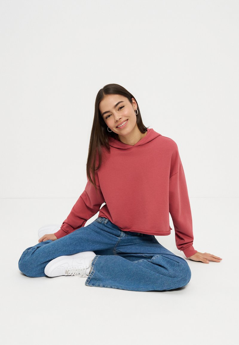 Joven mujer con sudadera roja, vaqueros azules y zapatillas blancas, sentada con las piernas cruzadas sobre un suelo blanco, sonriendo a la cámara.