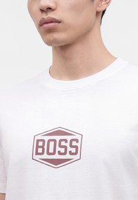 Νέος άντρας που φοράει λευκό T-shirt με λογότυπο εξάγωνο και τη λέξη "BOSS" τυπωμένη καφέ στο στήθος.
