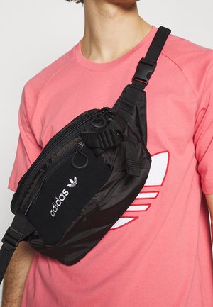 Mustassa nylonista valmistetussa olkalaukussa on useita vetoketjullisia lokeroita, säädettävä hihna ja valkoinen Adidas-logo. Laukkua on käytetty vaaleanpunaisen paidan päällä.
