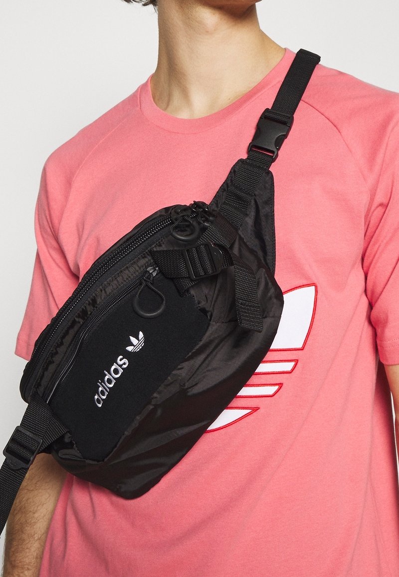 Černá crossbody taška vyrobená z nylonu, obsahující více zipových kapes, nastavitelný popruh a bílé logo Adidas. Nositá přes růžové tričko.