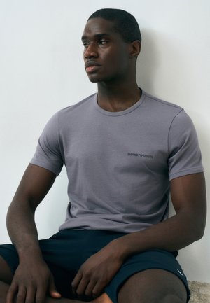 Junger Mann mit dunkler Haut, der ein graues Emporio Armani T-Shirt und schwarze Shorts trägt, sitzt mit überkreuzten Beinen an eine weiße Wand gelehnt.