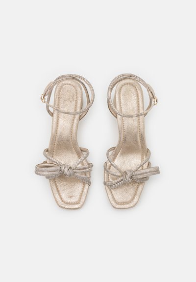 Sandales dorées pailletées avec un bout carré, présentant des brides croisées et un détail noué. Texture lisse avec un petit talon et une boucle à la cheville.