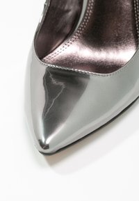 Chaussure à talon haut argentée à bout pointu avec finition brillante et doublure intérieure métallique sur fond blanc.