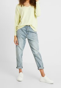 Jasnożółty, luźny sweter z dekoltem w serek, zestawiony z jasnoniebieskimi, lekko postrzępionymi jeansami z wysokim stanem oraz białymi trampkami.