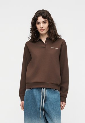 Femme aux cheveux bouclés foncés portant un sweat-shirt polo marron à manches longues et un jean bleu ample avec des cordons blancs.