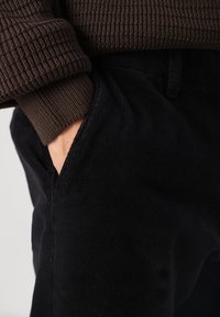 Pantaloni negri din catifea cu o suprafață texturată, având buzunare laterale. Sus este vizibil un pulover maro cu dungi, care acoperă parțial talia.