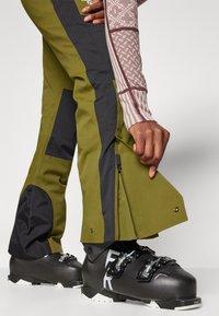 Skihosen in Olivgrün und Schwarz, mit Reißverschlussabschlüssen und verstärkten Knieflicken; getragen mit schwarzen Skischuhen, die metallische Akzente präsentieren.