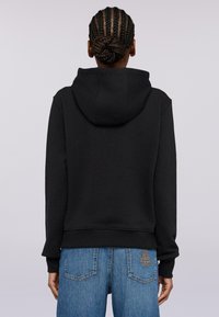 Svart huvtröja med dragsko och ribbad nederdel, tillverkad av mjukt tyg. Modellen har den på sig tillsammans med blå jeans med detaljer vid bakfickan.