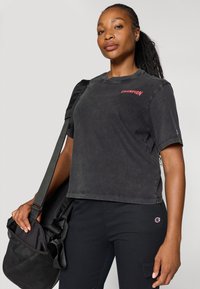 Sort t-shirt med korte ærmer har en afslappet pasform og et fremtrædende rødt "CHAMPION" logo på brystet. Modellen holder en sort skuldertaske.