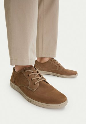 Person trägt beige Hose und braune legere Schnürschuhe mit hellen Sohlen, steht vor weißem Hintergrund.