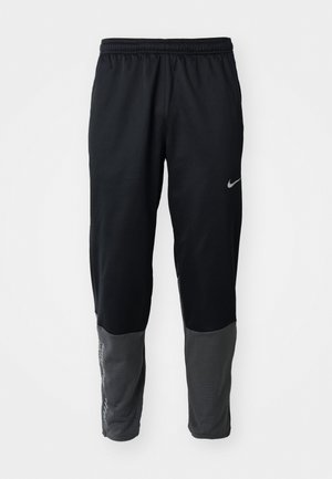 Melni un tumši pelēki sporta bikses ar elastīgu jostu, kurām augšējā kreisajā augšstilbā un apakšējā kreisajā kājā ir Nike logo.