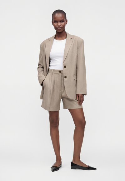 Femme portant un blazer beige oversize et un short assorti avec un t-shirt blanc et des ballerines noires pointues, debout avec une main dans la poche.