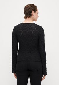 Czarny top z długim rękawem o fakturowanym, kwiatowym wzorze. Dopasowany krój, okrągły dekolt i teksturowana tkanina. Noszony z czarnymi spodniami.