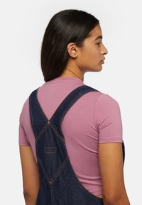 Denim overall in dark wash with gold stitching, worn over a light pink, short-sleeve t-shirt. Features cross-back straps and a side pocket.