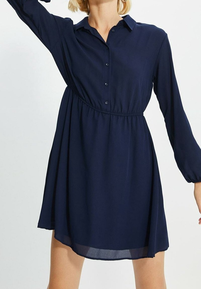 Femme portant une robe bleu marine à manches longues, boutonnée, avec une taille cintrée et un col, visible de mi-cuisse jusqu'au cou.