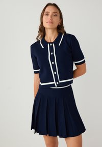 Twinset di un blu navy a coste con maniche corte, rifinitura bianca, chiusura con bottoni e gonna plissettata. Il tessuto ha una finitura strutturata.