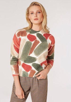 Kolorowy, teksturowany sweter z dzianiny z abstrakcyjnymi wzorami w odcieniach zieleni, czerwieni i beżu, zestawiony z w kratkę spodniami. Okrągły dekolt, długie rękawy.