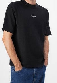 Black cotton t-shirt with a crew neck, short sleeves, and a small white "Selected" logo on the chest. Worn with blue denim jeans.