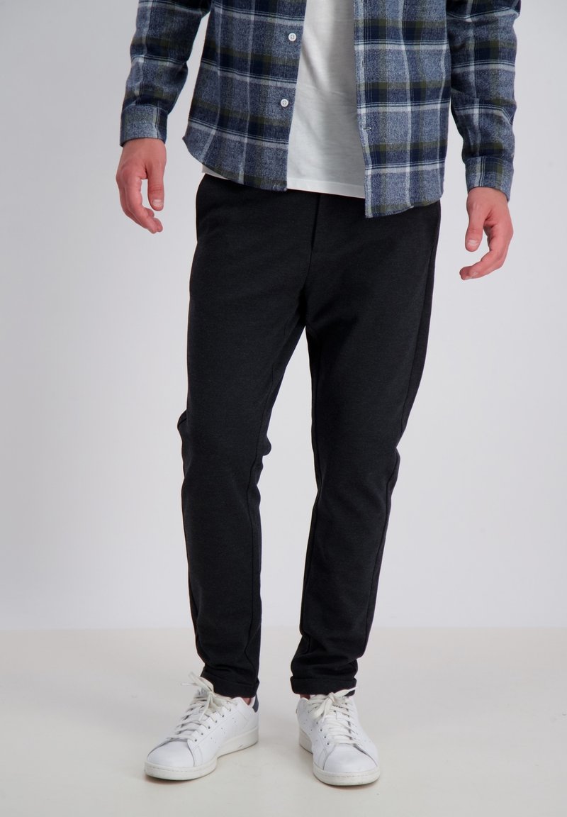 Schwarze Jogginghose aus einem weichen Material, mit elastischem Bund und schmal zulaufenden Beinen, kombiniert mit einem blauen Karoshirt und weißen Turnschuhen.