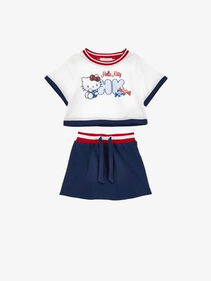 Fehér, rövidített póló piros és tengerészkék szegéllyel, Hello Kitty grafikával és cukorkákkal, tengerészkék szoknyával párosítva, piros-fehér csíkos derékpánttal.