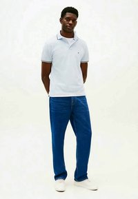 Jovem de pé com as mãos atrás das costas, vestindo uma camisa polo azul clara, jeans azuis e ténis brancos, em um fundo simples.