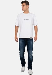 Uomo che indossa una camicia bianca a maniche corte con un piccolo logo, jeans blu scuro e scarpe da ginnastica bianche, in piedi e che guarda di lato.