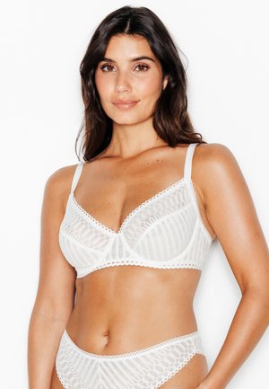 Femme aux longs cheveux foncés portant un soutien-gorge en dentelle blanche et des sous-vêtements assortis sur un fond blanc uni.