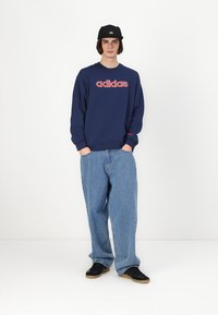 Mornarska dukserica s crvenim "adidas" logotipom, uparena s labavim svijetloplavim trapericama i crnim tenisicama s gumenim potplatom; nošena s crnom kapom.