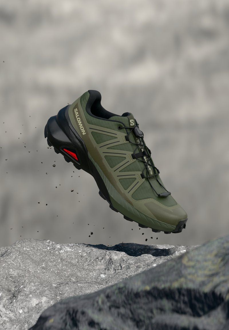 Scarpa da trail running Salomon verde e nera a mezz'aria sopra un terreno roccioso con particelle di sporco che cadono dalla suola.