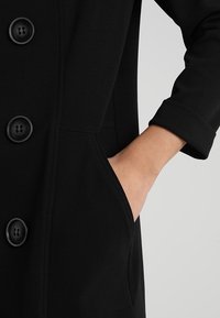 Černý kabát s bezšvým designem, vybavený velkými černými knoflíky, bočními kapsami a rukávy s ohrnutými manžetami. Vyrobený z hladké látky.