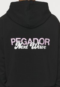 Čierna mikina s kapucňou, na chrbte s tučným nápisom "PEGADOR Next Wave" v ružovej a bielej farbe; vyrobená z mäkkej, hrubej tkaniny.