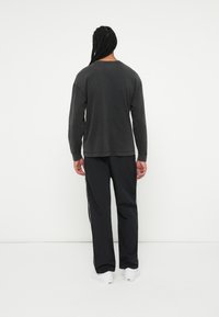 Crna majica dugih rukava s opuštenim fitom, u paru s crnim trenirka hlačama koje imaju bijele bočne pruge i bijelim sportskim cipelama.