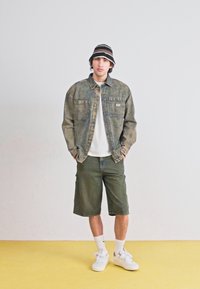 Giacca di jeans in verde sbiadito con due tasche sul petto, abbinata a pantaloni corti verde oliva. Maglietta bianca sotto e cappello a secchiello a righe nere. Sneakers bianche.