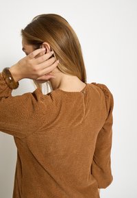 Hnedý textúrovaný top s balónovými rukávmi a okrúhlym výstrihom so zapínaním na malý gombík v zadnej časti. Hladká, rebrovaná tkanina.