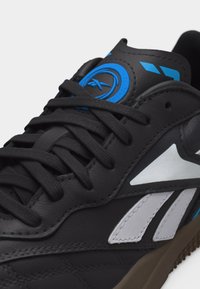 Μαύρο αθλητικό παπούτσι με μπλε κυκλικό λογότυπο στη γλώσσα, λευκές πλευρικές λεπτομέρειες και μαύρα κορδόνια σε καφέ σόλα, σε λευκό φόντο.