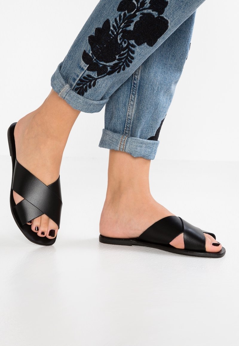 Pieds chaussés de sandales en cuir noir à brides croisées et bouts ouverts, associées à un jean bleu retroussé avec une broderie florale noire.