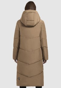 Cilvēks, kura mugurā ir garš, brūns, quilted puffer mētelis ar lielu kapuci, redzams no mugurpuses uz vienkārša fona.