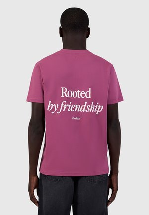 Man met een roze t-shirt waarop "Rooted by friendship" in het wit op de achterkant gedrukt staat, staande tegen een effen lichte achtergrond.