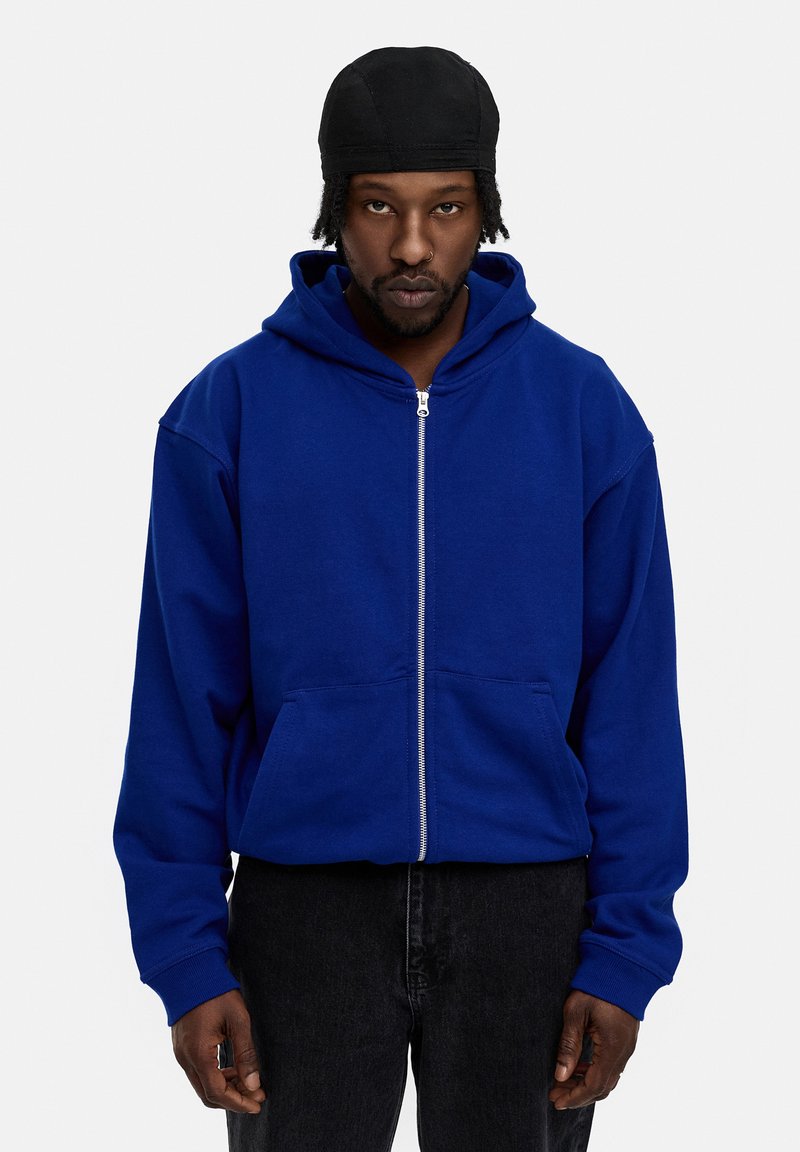 Blaue Kapuzenjacke mit Reißverschluss und vorderen Taschen, aus weichem Material gefertigt. Verfügt über eine große Kapuze und gerippte Bündchen, getragen mit dunklen Jeans.