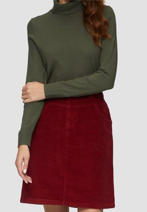 Femme portant un pull à col roulé vert olive et une jupe en velours côtelé rouge taille haute avec des poches, debout devant un fond uni.