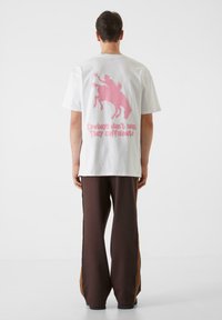 Biela bavlnená tričko s ružovou grafikou kovboja na koni a textom. Skombinované s hnedými nohavicami so bočnými pruhmi.